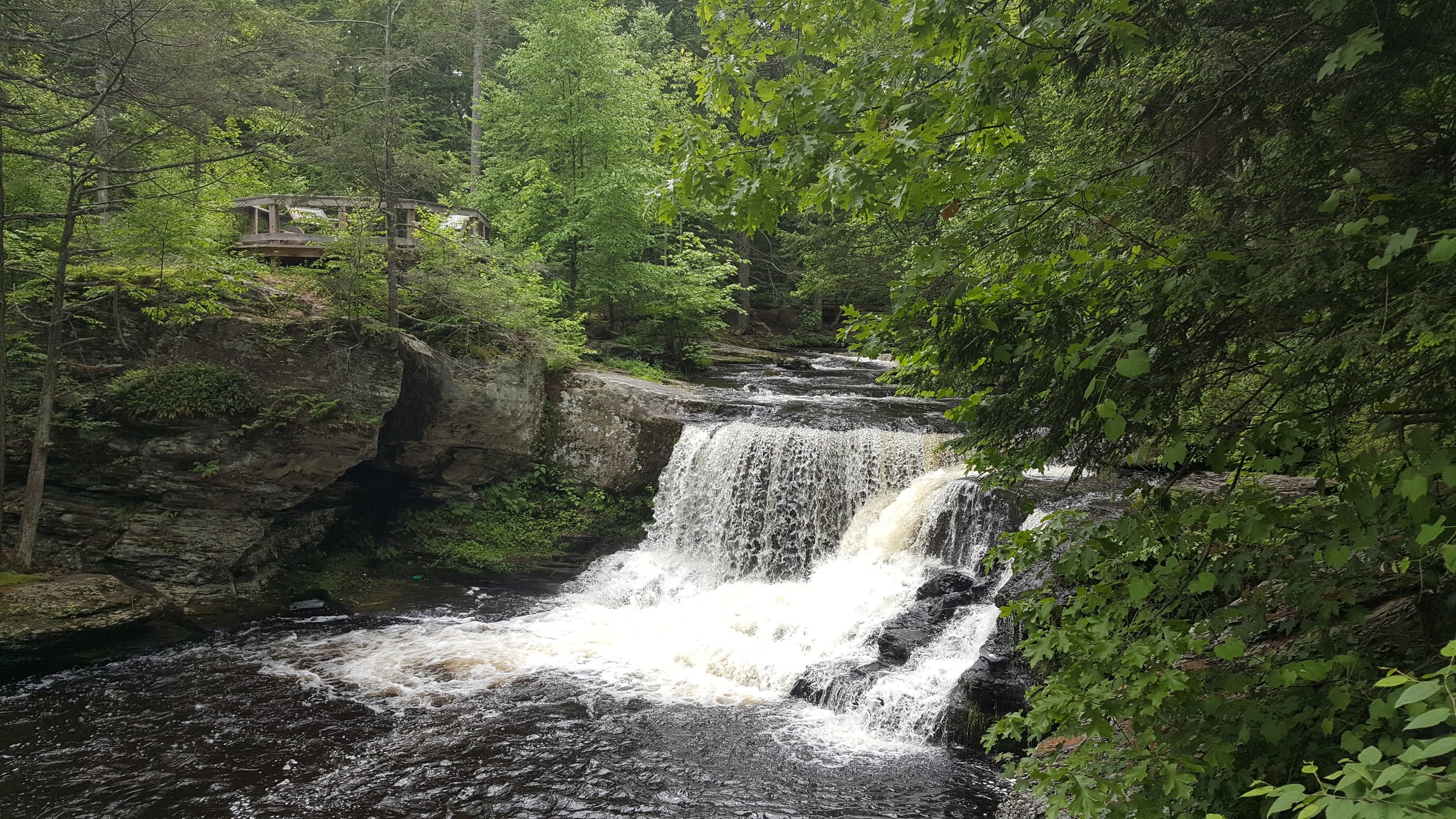 Wide view of a waterfall spilling over a rock ledge with a wooden overlook among the trees.